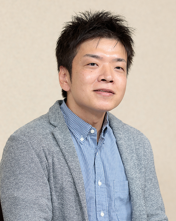 A portrait photograph of a male speaker from Nippon Paint Holdings, wearing a blue check shirt and grey blazer, facing forward with a neutral background.