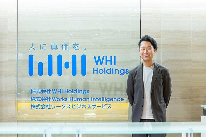 A man in business attire stands smiling in the reception area of WHI Holdings. The company logo and names of affiliated companies, including Works Human Intelligence and ワークスビジネスサービス, are prominently displayed on a glass wall in Japanese and English.