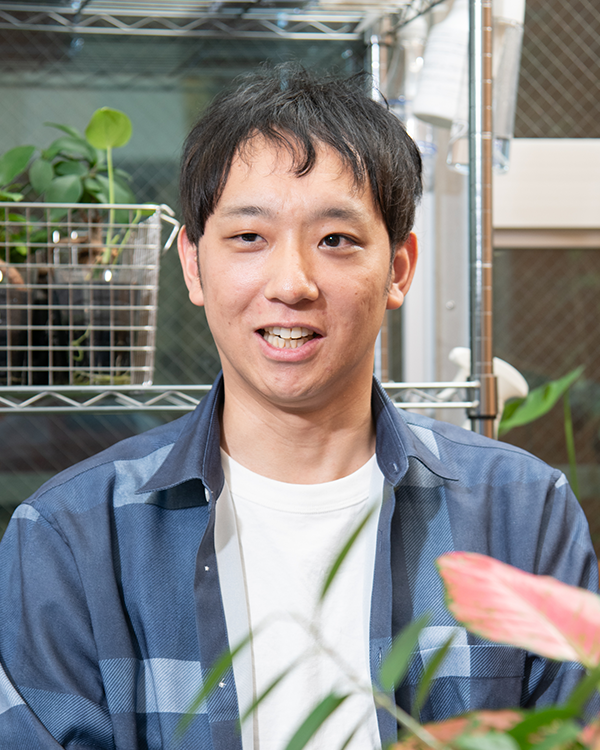 A portrait of a person with short dark hair, wearing a blue shirt and white t-shirt, sitting in front of shelves with green plants and baskets, with additional plants in the foreground.