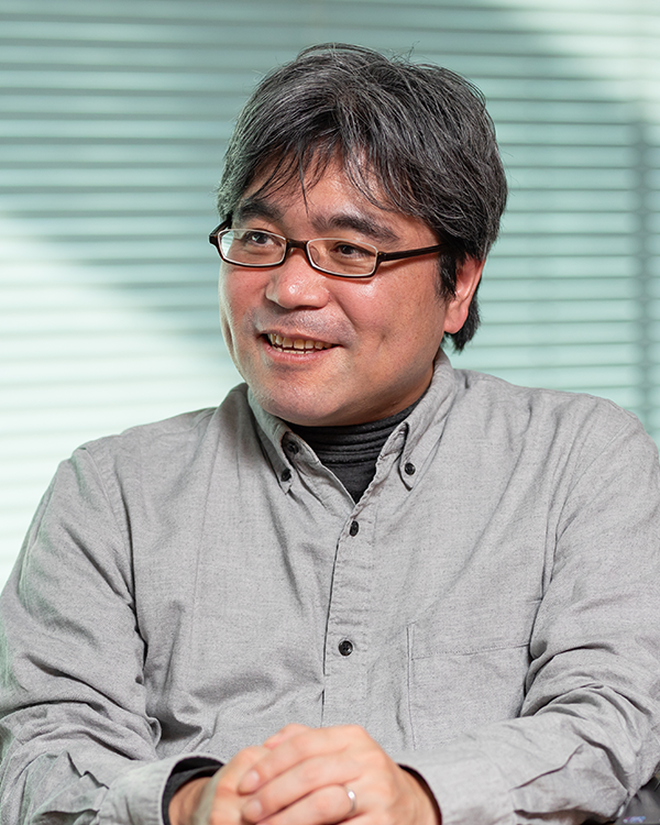 Portrait of a speaker featured in the Bandai Namco case study, sitting and smiling, wearing glasses and a grey shirt, with a blurred background of horizontal blinds.