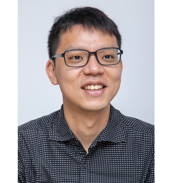 A portrait photo of an individual wearing glasses and a black patterned shirt, smiling in front of a light gray background.