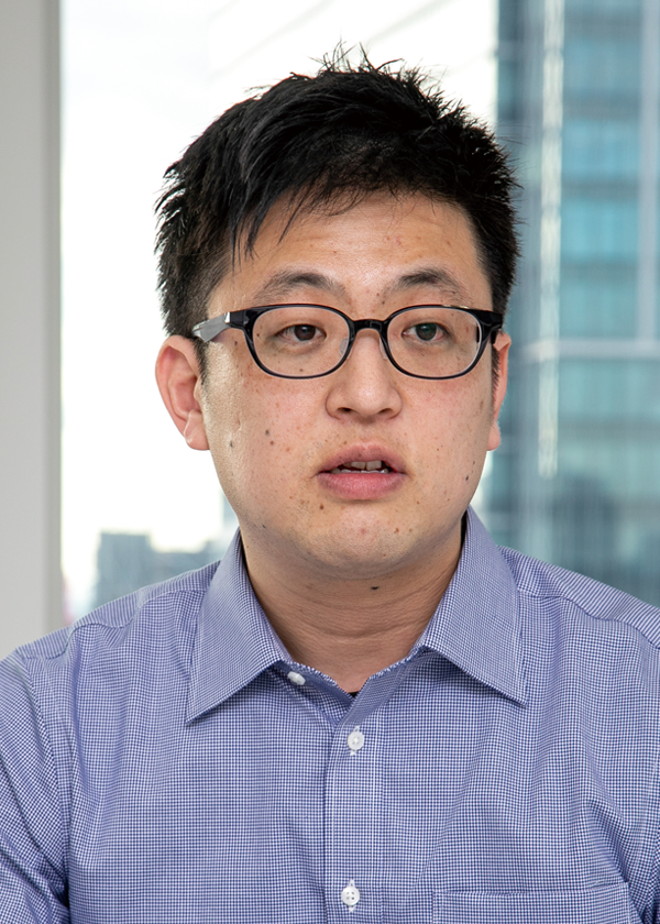 Business portrait of a person wearing glasses and a collared shirt, photographed in an office setting with windows and city buildings in the background.