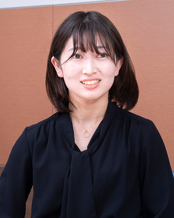 A person wearing a black blouse and a simple gold necklace with a circular pendant, seated against a brown background.