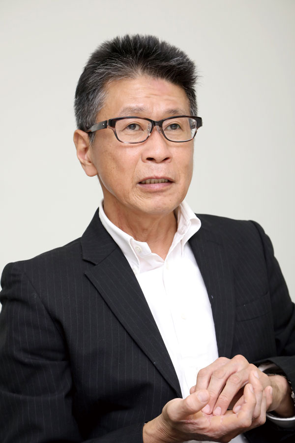 A man wearing glasses and a dark suit with a white shirt, sitting and gesturing with his hands, in front of a plain background.