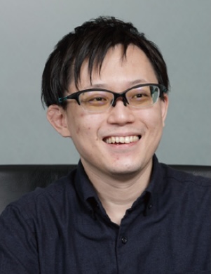 A person with short dark hair wearing glasses and a dark shirt, smiling while looking slightly to the side.