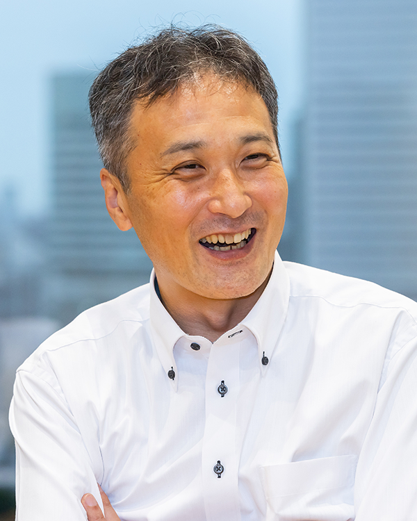 A smiling executive in a white shirt, seated indoors with a cityscape in the blurred background.