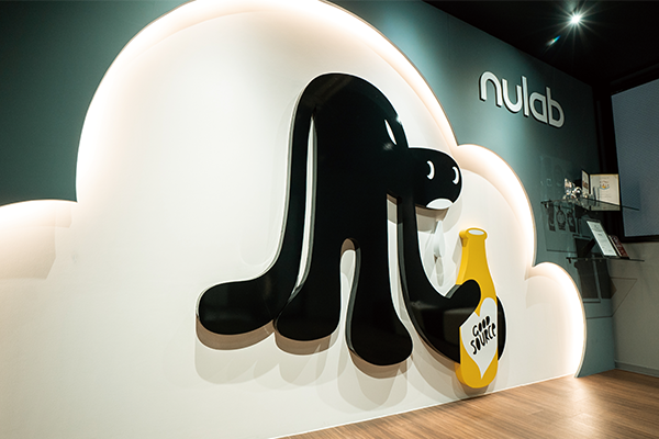 A photo of the Nulab office featuring a wall decoration with a stylized black character holding a yellow sauce bottle, set against a white cloud-shaped background and the 'nulab' logo on the wall.