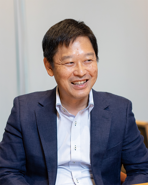 A smiling person wearing a white shirt and a dark business suit jacket, seated in an office or meeting room setting.