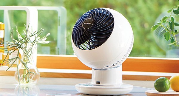 A white Iris Ohyama circulator fan is placed on a kitchen counter near a window, with plants and fruit nearby.
