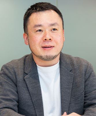 Portrait of a male speaker from V-Cube featured in an AWS solution case study, dressed in a gray blazer and white shirt, seated and speaking.