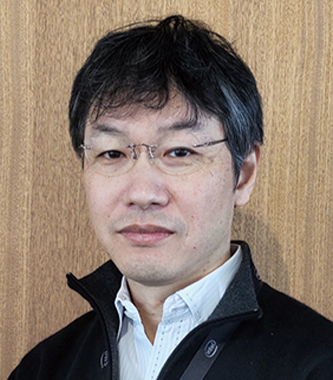 A portrait of a man wearing glasses, standing in front of a wooden background. He is wearing a white shirt and a dark jacket. (Konica Minolta case study asset)