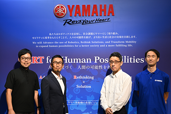Alt-text: Four individuals stand in front of a Yamaha promotional backdrop featuring the slogan "ART for Human Possibilities" and text about robotics and mobility solutions.