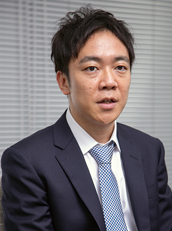 A business portrait of a man in a navy suit and patterned tie, seated in front of a window with blinds.