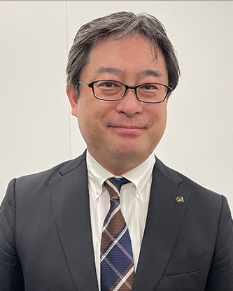 A portrait of a business professional wearing a suit, tie, and glasses, standing in front of a plain background.