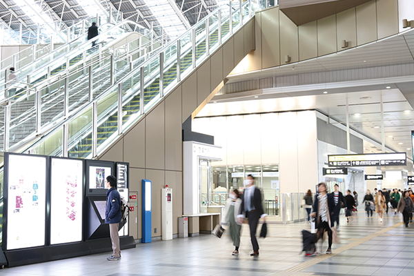 A modern train station interior with people walking, a man using an information kiosk, and escalators in the background.