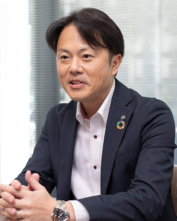 A businessman in a dark suit and white shirt is being interviewed. He is wearing a watch and a colorful SDGs (Sustainable Development Goals) pin on his lapel, seated indoors with a blurred office background.