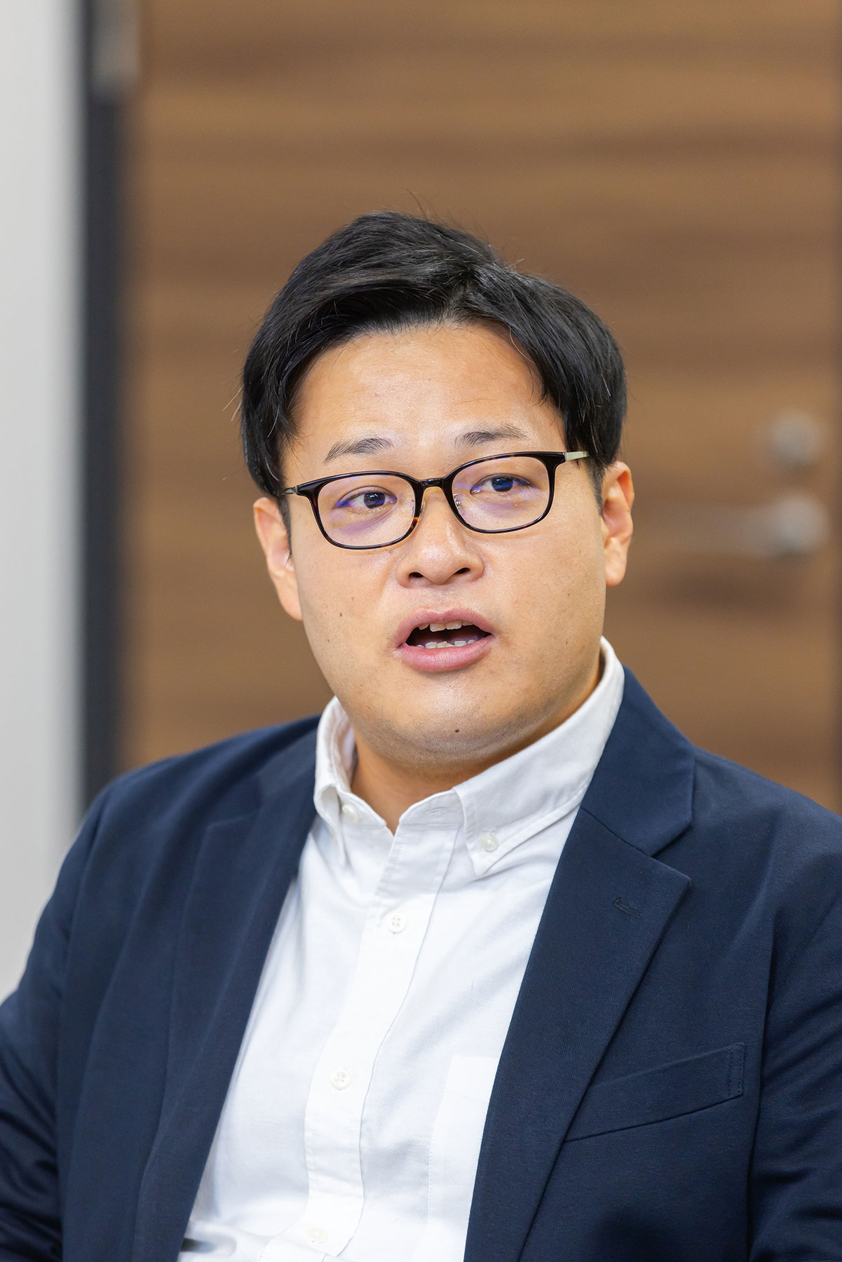 A person wearing a white collared shirt and navy blazer, seated indoors with a wooden door in the background.