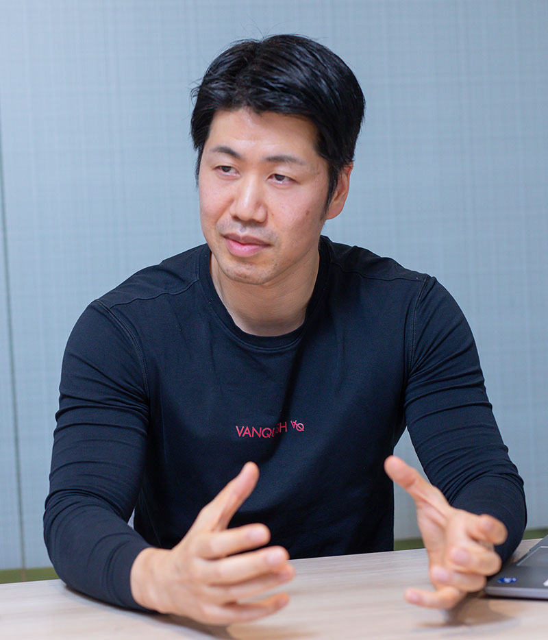 A person wearing a black long-sleeve shirt with "VANQUISH" text, gesturing with their hands while seated at a table.