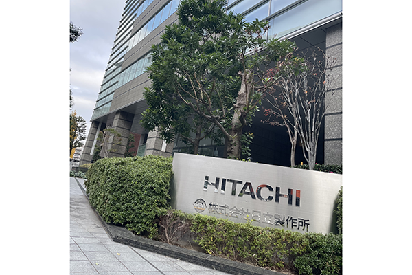 Photograph of the Hitachi headquarters building sign in Japan, featuring the company name and logo mounted on a stone plaque outside the building with trees and a modern office exterior in the background.