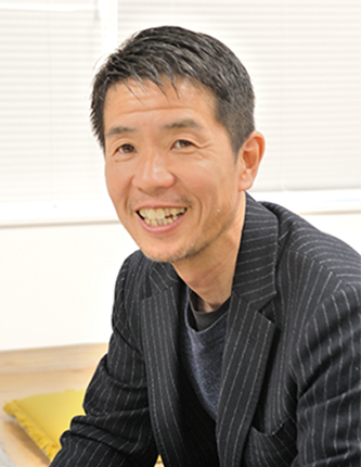 A smiling man wearing a dark striped blazer, seated in an office setting with light-colored blinds in the background.