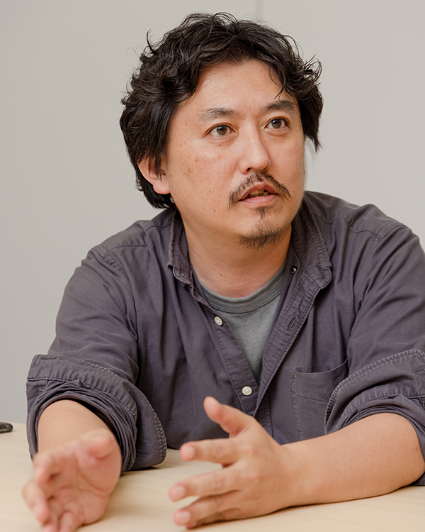 A man with dark hair and a goatee, wearing a dark button-up shirt, sits at a table and gestures with his hands during a discussion or interview.