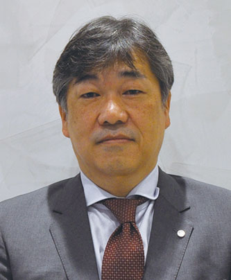 A person wearing a gray suit, white shirt, and patterned red tie, standing against a light gray background.