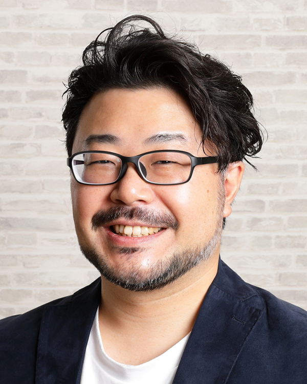 A person wearing a navy blazer and white shirt against a light brick wall background.