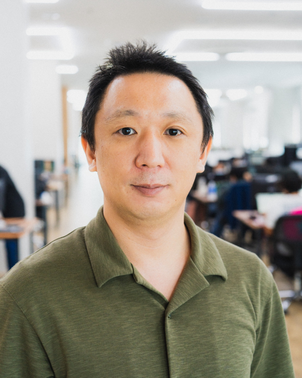 A person wearing a green collared shirt standing in a bright, modern office space with desks and people working in the background.