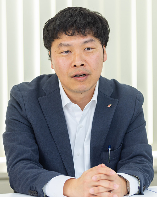Portrait of a man in a suit from the ecbeing case study, sitting with hands folded, photographed in front of vertical blinds.