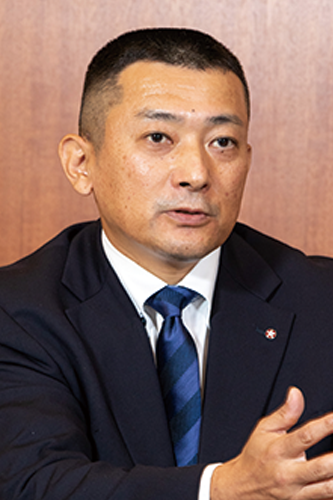 A portrait of Mr. Kuroki of Nihon Shokken, dressed in a suit and tie, speaking in a business setting. Background features a wooden wall.