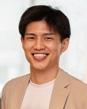 A smiling man wearing a light-colored business casual jacket and white shirt, standing indoors.