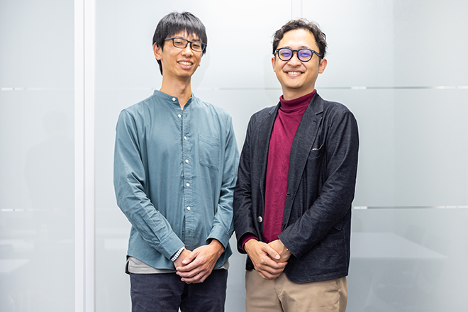 Two men standing and smiling in an office environment. This image is used for the Hitachi Group case study.