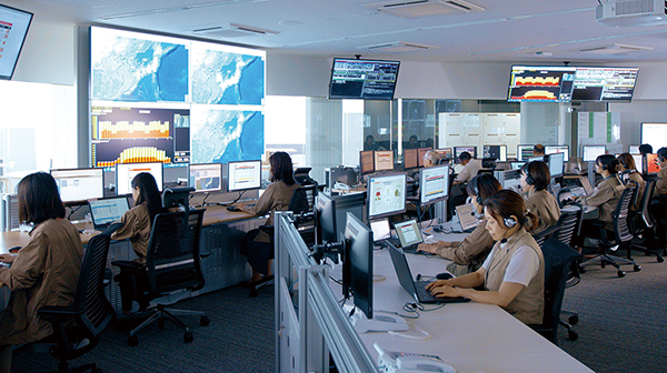 A modern control room with multiple operators working at computer stations. Large display screens show maps and data charts, illustrating a high-tech environment for monitoring energy systems, as featured in the Yanmar Energy Systems AWS case study.