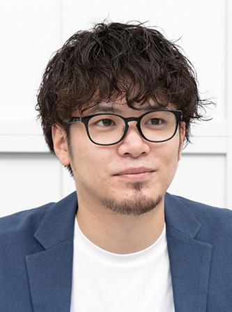 Portrait of a man with curly hair, wearing glasses, a blue blazer, and a white shirt, seated in front of a light background.
