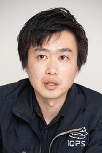 Portrait of an iQPS employee wearing a jacket with the iQPS logo, neutral background.