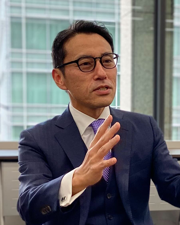 A business leader wearing a blue suit and glasses, gesturing while speaking during an interview in an office setting with large windows and a city view in the background.