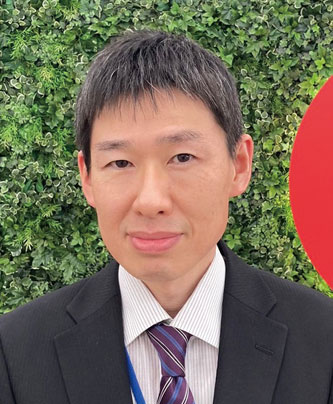 A person in formal attire standing in front of a green leafy background with a partial red circle on the right.