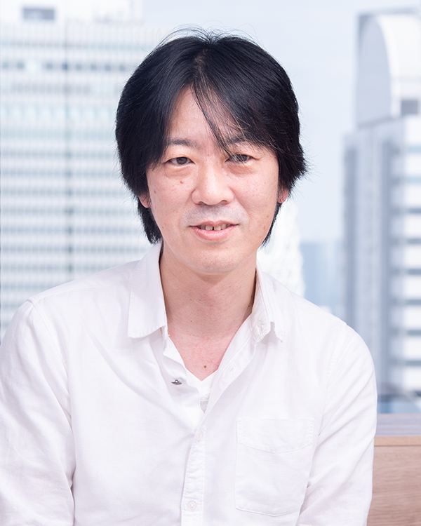 A portrait photo of a man in a white shirt, posed indoors with an urban cityscape in the background. Used for the RIZAP case study featuring Yoshinori Nagashima.