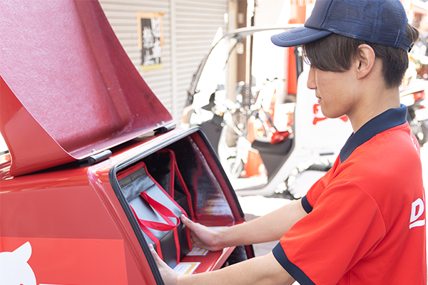 A Demae-can delivery staff member in uniform is loading a food container into a red delivery vehicle, preparing for deliveries.
