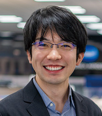 A professional portrait of a businessperson in an office environment, associated with Konica Minolta, wearing glasses and a suit, smiling at the camera.