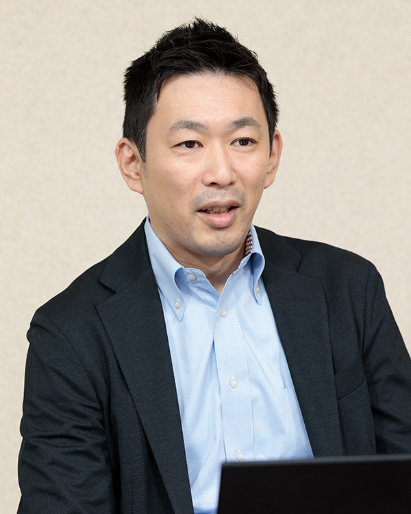 A person wearing a light blue button-up shirt and a dark blazer, sitting in front of a laptop.