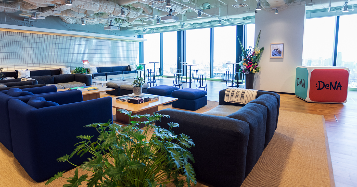 A spacious and modern lounge area in the DeNA office, featuring contemporary sofas, tables, plants, and large windows with a city view.