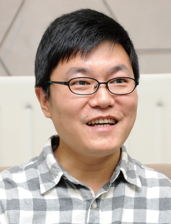 A smiling man wearing glasses and a checkered shirt, seated indoors.