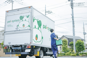 A Pal System delivery truck with branding and a delivery worker carrying a container, photographed in a residential neighborhood.