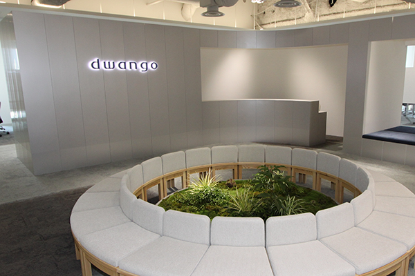 The image shows a modern office lobby at Dwango, featuring a circular indoor garden surrounded by cushioned seating. The design creates a relaxing and inviting environment in the reception area.
