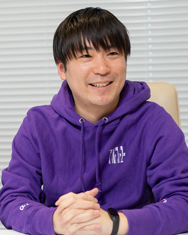 Person wearing a purple hoodie and a black wristwatch, seated at a table with hands clasped.