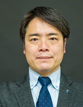 A person wearing a gray suit jacket, white shirt, and blue polka-dot tie against a dark background.