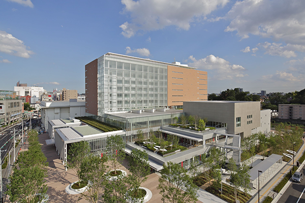 Modern multi-story building with glass and brick facade, surrounded by landscaped greenery and pathways under a partly cloudy sky.