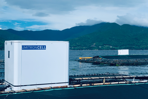 A photo of the UMITRON CELL device used in aquaculture, shown on a fish farming platform with water and mountains in the background under a cloudy sky.