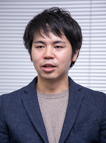 A portrait of a business professional wearing a navy blazer and beige sweater, standing in front of window blinds.